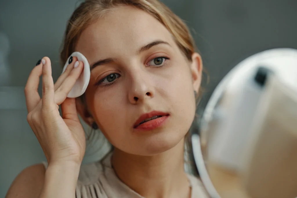 woman cleansing face with cotton pad at mirror